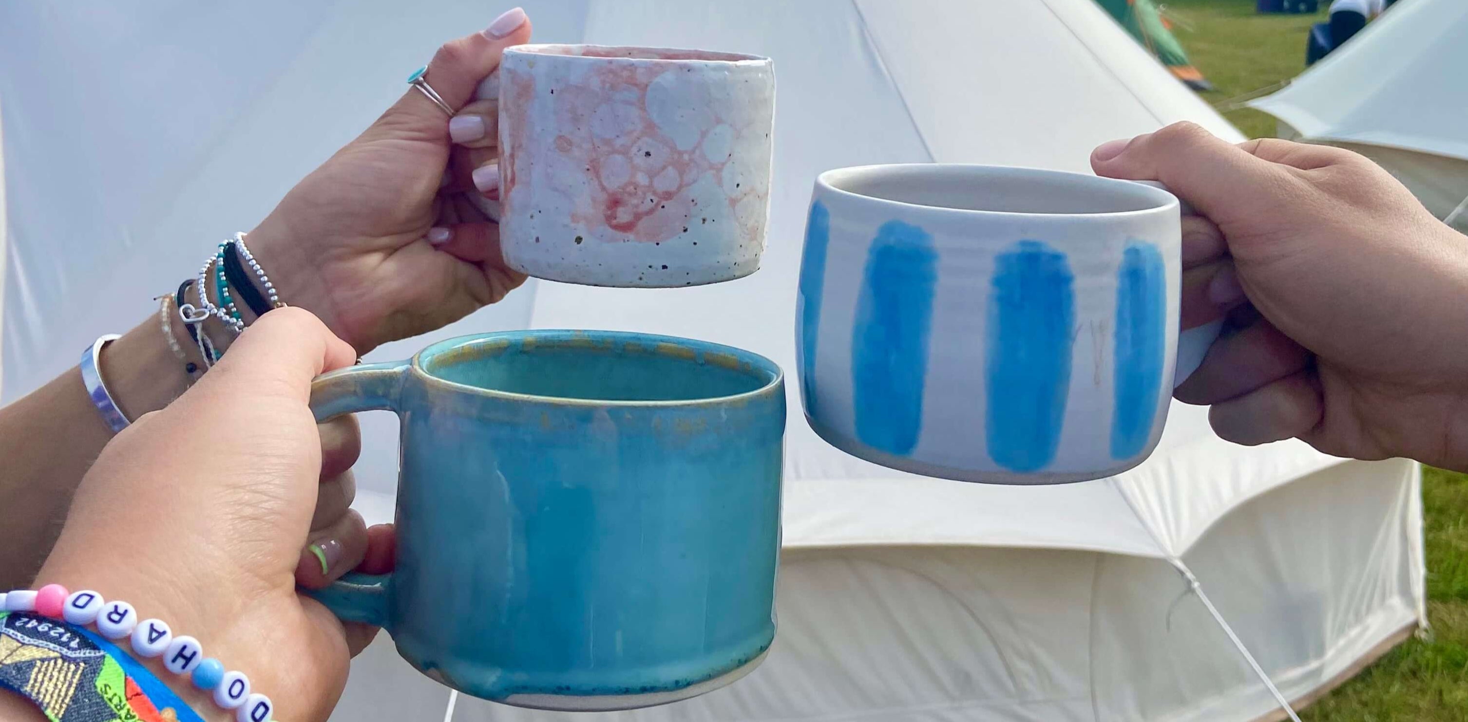 Three ceramic mugs with different designs held by hands against a tent background