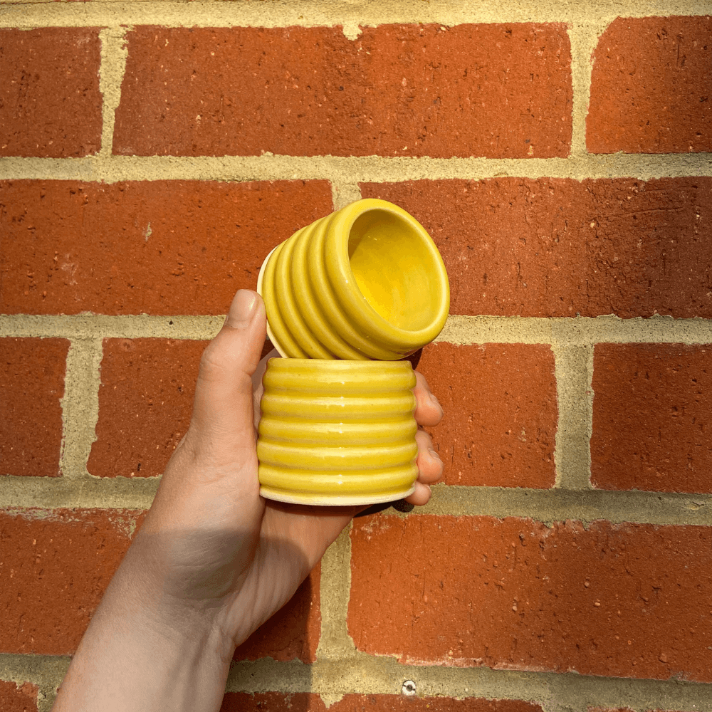 Hand holding two yellow ceramic espresso cups against a brick wall